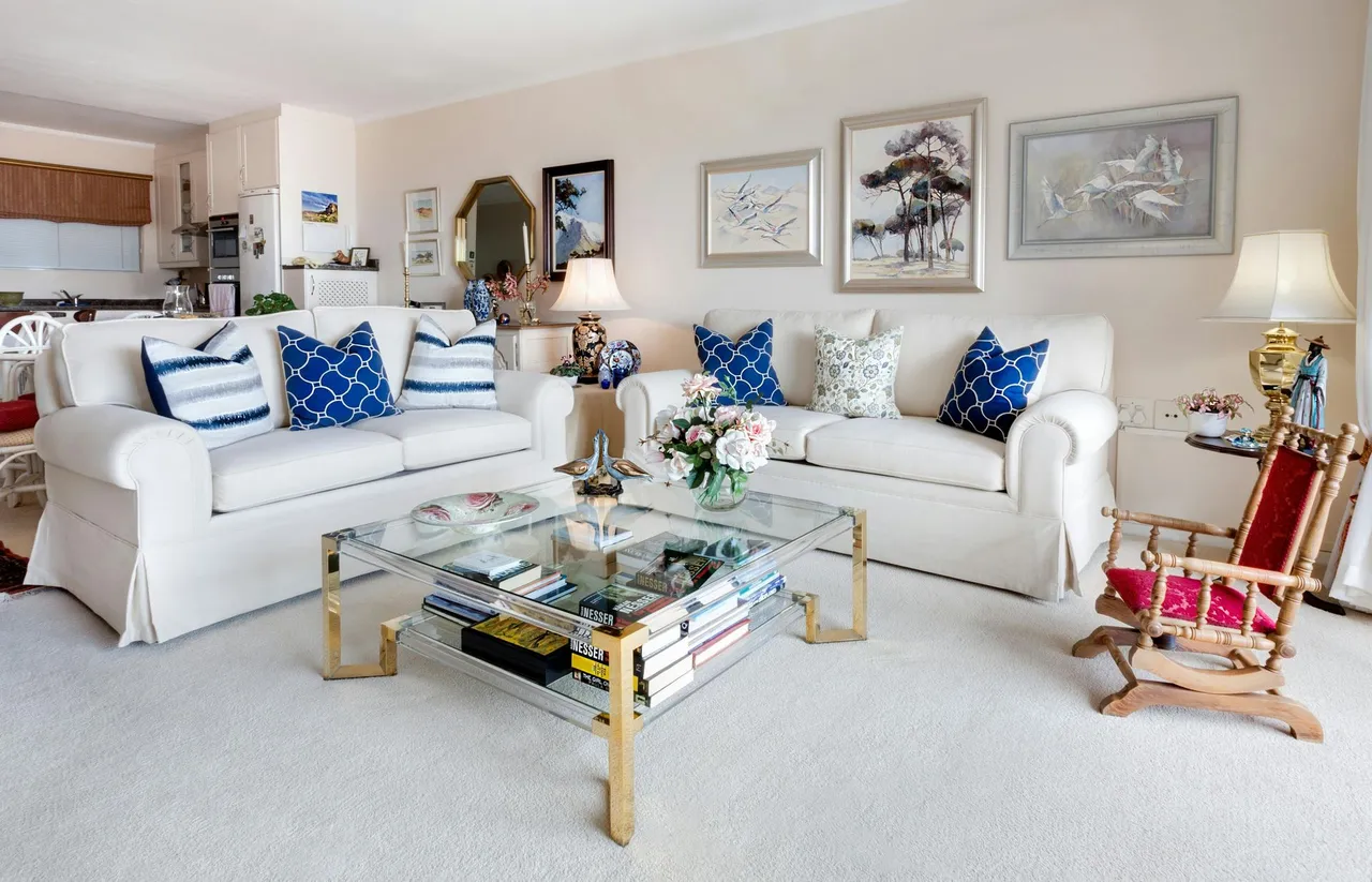 Bright white living room with two white sofas, blue pillows, a glass coffee table, and framed wall art