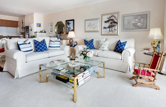 Bright white living room with two white sofas, blue pillows, a glass coffee table, and framed wall art