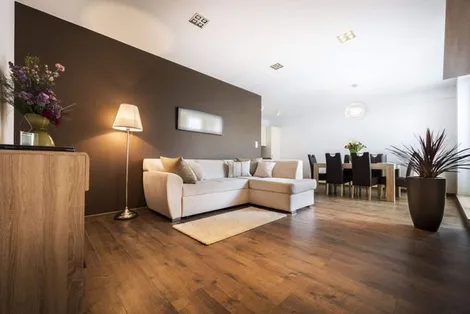 Modern living room with a brown accent wall, beige sectional sofa, floor lamp, and a dining area in the background.