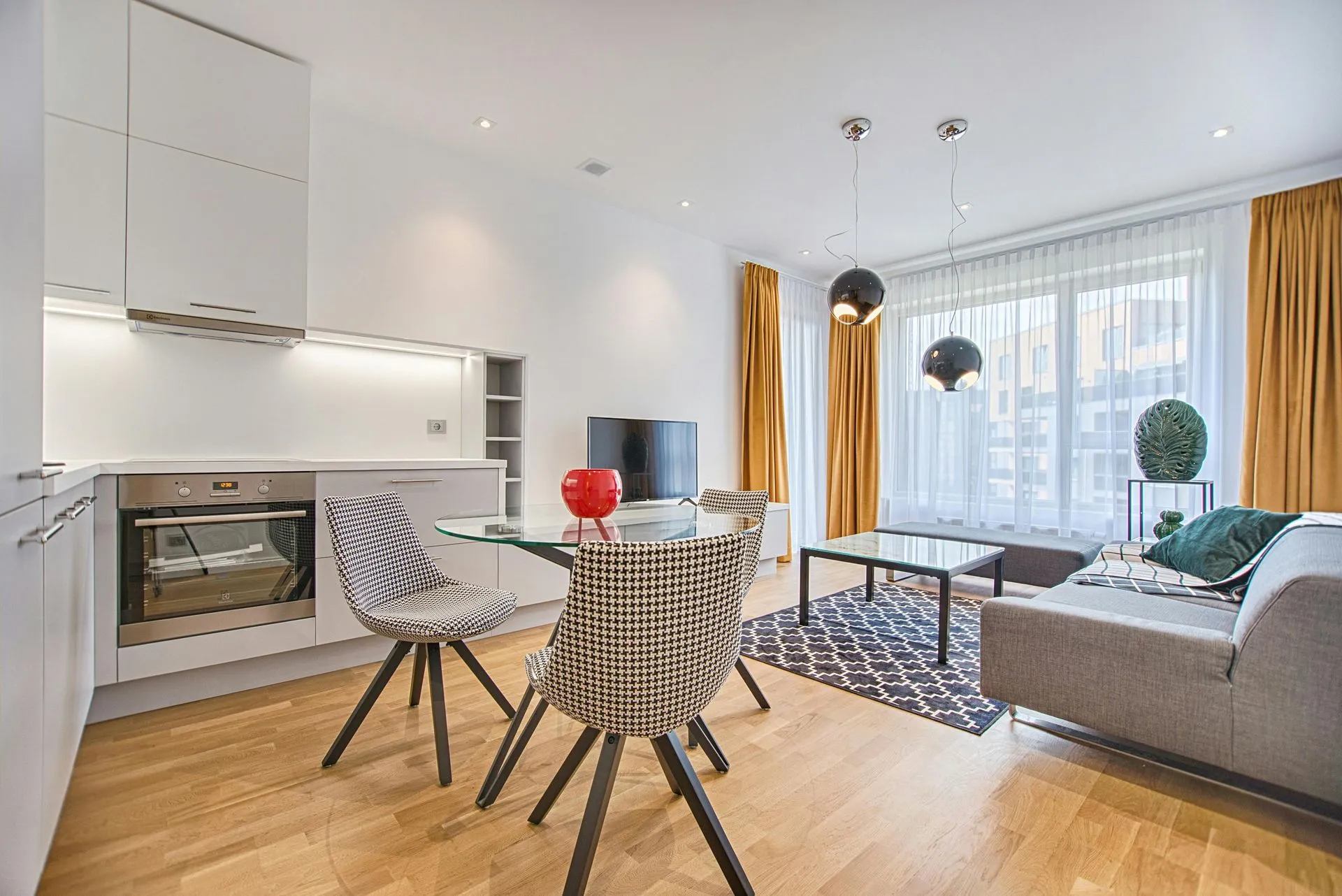 A modern living room with hardwood floors, a dark sofa, a coffee table, and large windows overlooking autumn trees.