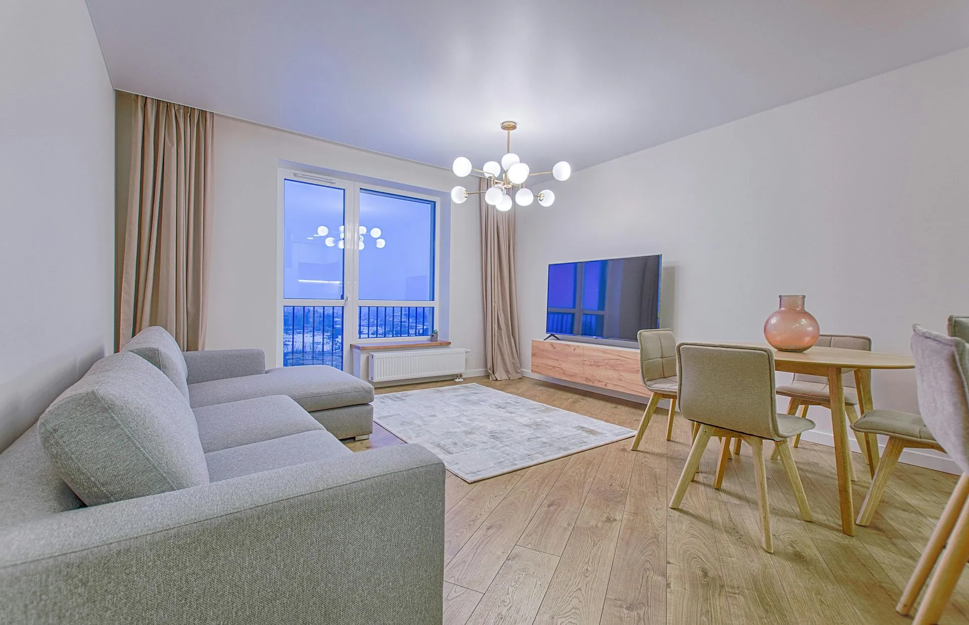 Bright modern living room with gray sofa, dining table, TV, and balcony view at duskD