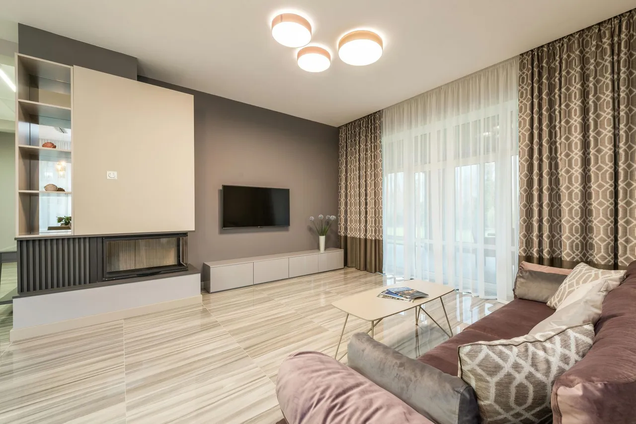 A modern living room with hardwood floors, a dark sofa, a coffee table, and large windows overlooking autumn trees.